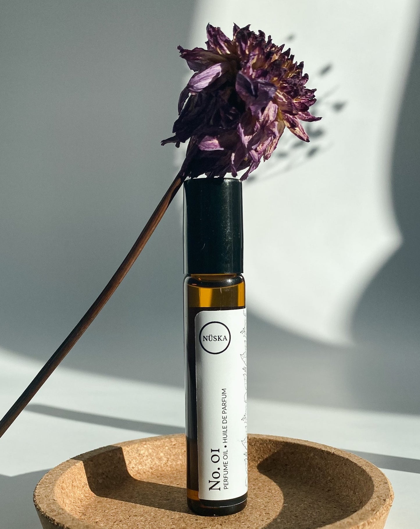 Nuska Botanical Perfume Oil No.1 in a roll-on amber glass bottle, surrounded by natural light and a dried purple flower, highlighting its floral and citrus scent.