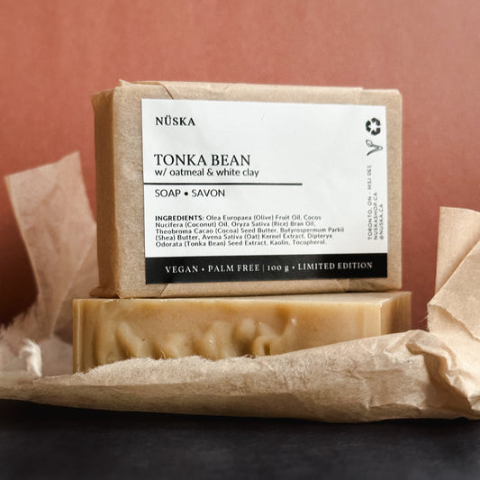A rectangular bar of Nuska Tonka Bean with Oatmeal and white clay natural soap with a paper package and minimal label on top of another soap bar package free.