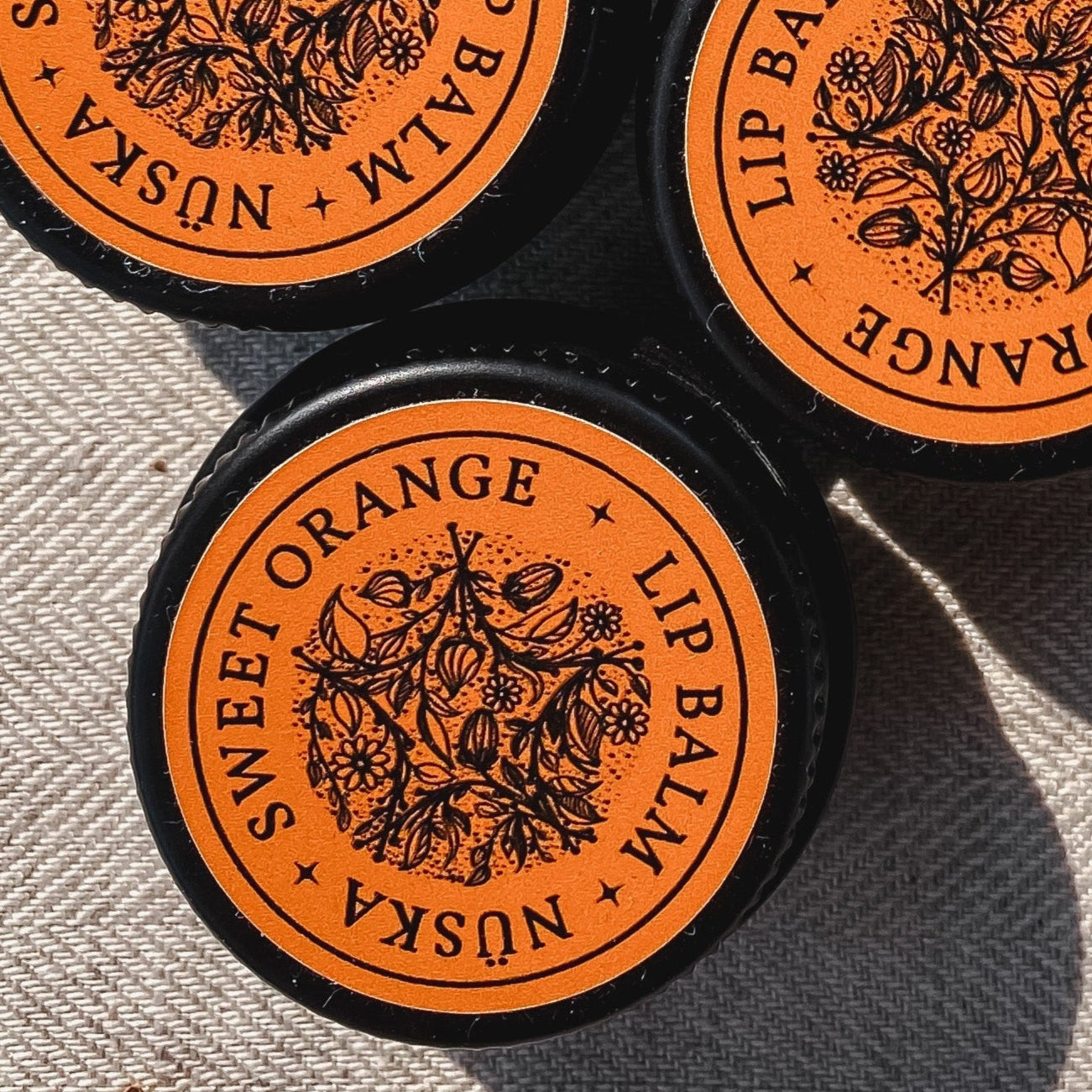 A black aluminum tin with an orange label of Nuska sweet orange natural lip balm.