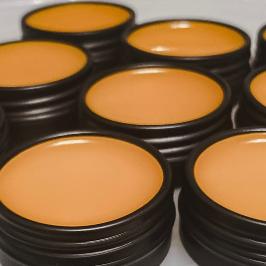 An open black aluminum tin of Nuska Sweet Orange Natural Lip Balm, showing the creamy texture and orange color of the moisturizing balm.