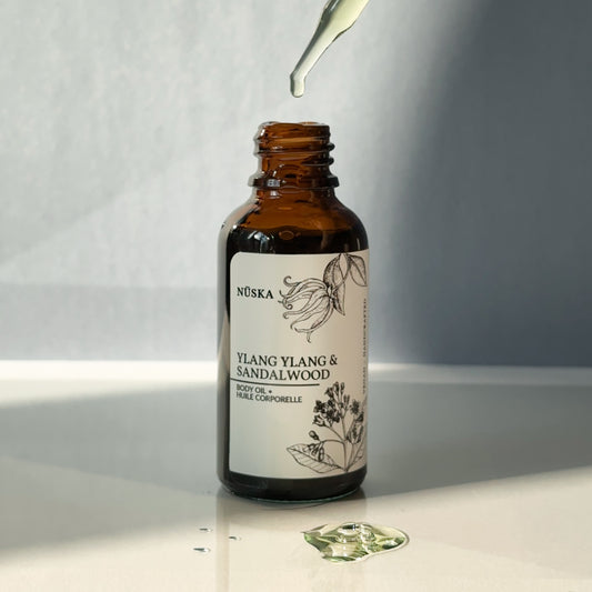 Travel size Nuska Ylang Ylang & Sandalwood Botanical body oil in an amber glass dropper bottle and minimal label, with a drop of body oil in the front and blue background.