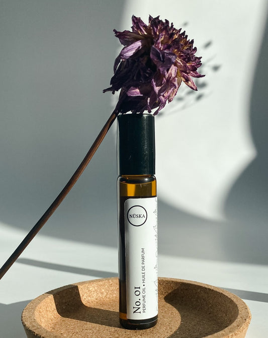 Nuska Botanical Perfume Oil No.1 in a roll-on amber glass bottle, surrounded by natural light and a dried purple flower, highlighting its floral and citrus scent.
