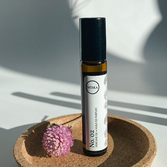 Nuska Botanical Perfume Oil No.2 in a roll-on amber glass bottle, surrounded by natural light and a dried pink flower, highlighting its oriental scent from essential oils.