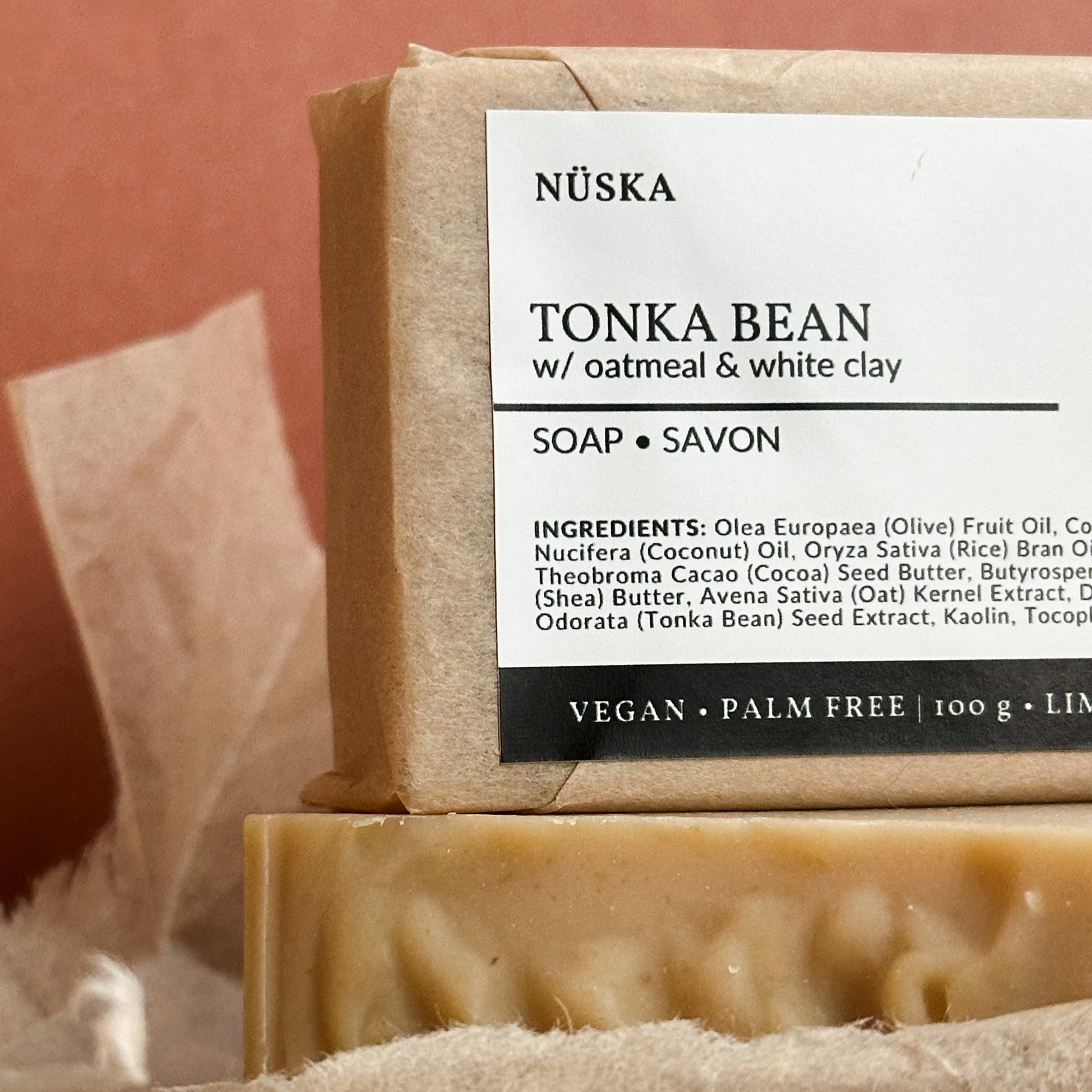 A close up view of a rectangular bar of Nuska Tonka Bean with Oatmeal and white clay natural soap with a paper package and minimal label on top of another soap bar package free.