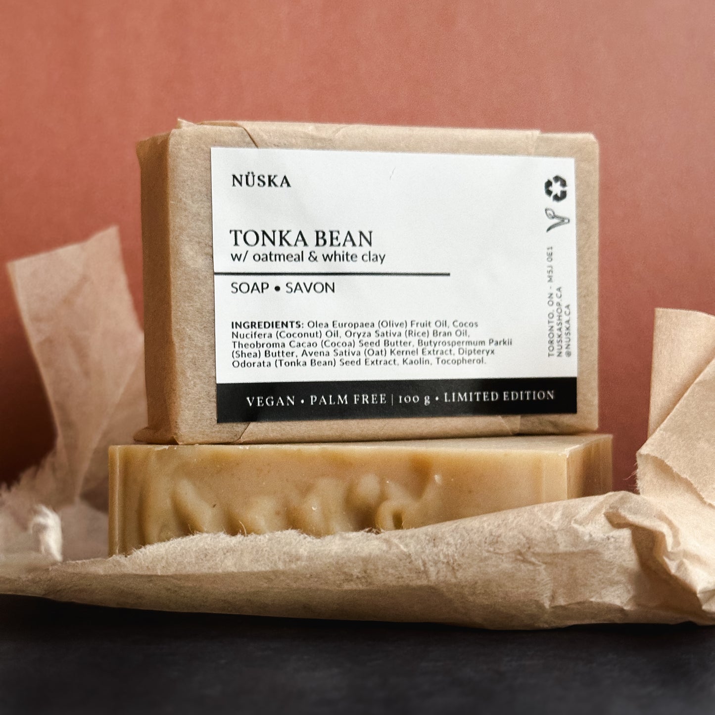 A rectangular bar of Nuska Tonka Bean with Oatmeal and white clay natural soap with a paper package and minimal label on top of another soap bar package free.