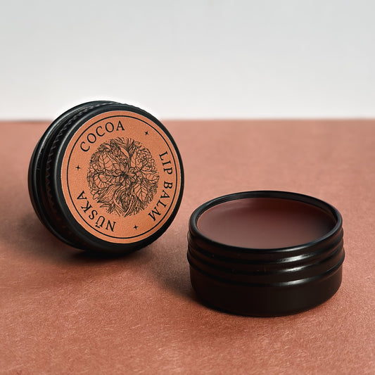 A black aluminum tin with a brown label of Nuska cocoa natural tinted lip balm, and another open tin showing the colour and texture of the lip balm.