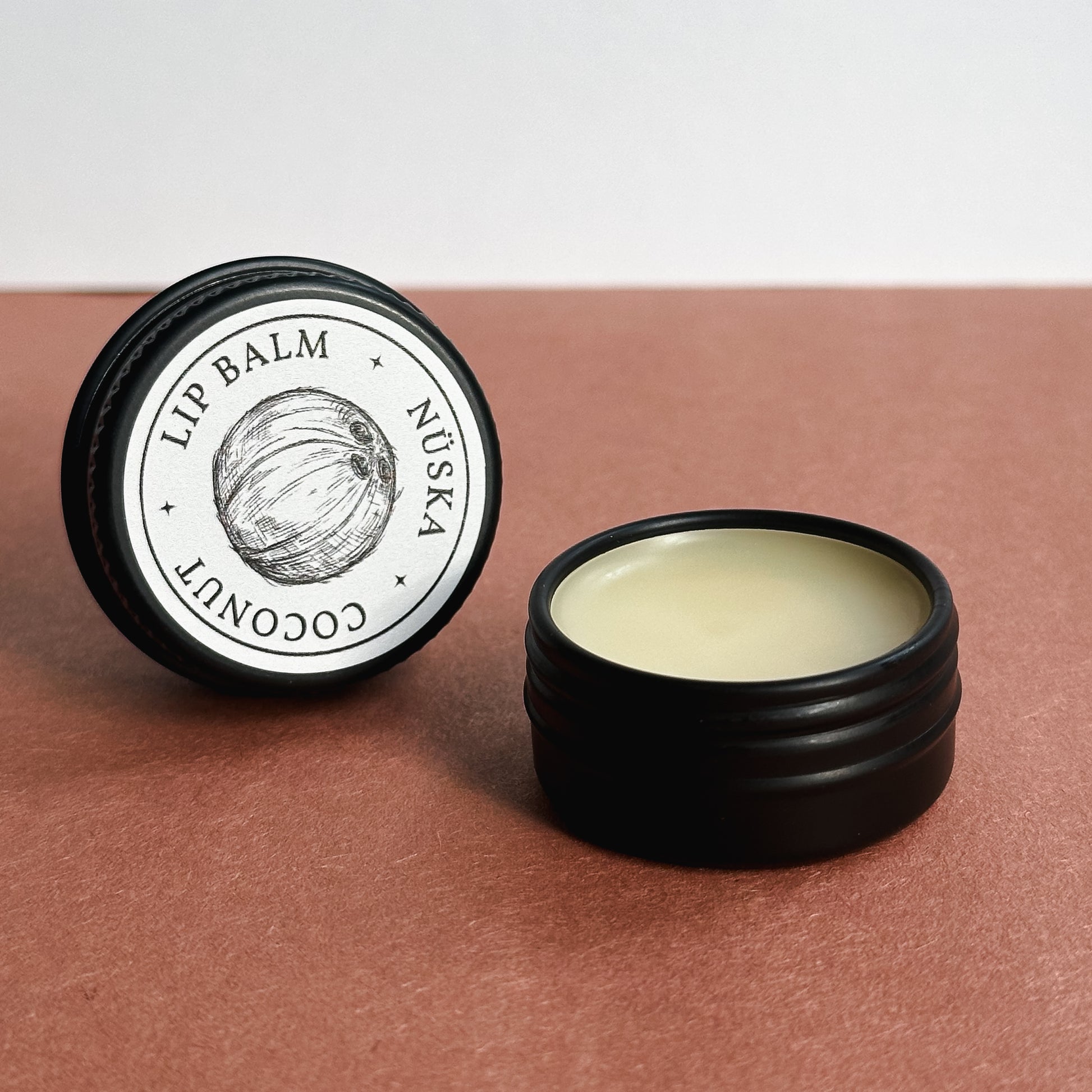 A black aluminum tin with a gray label of Nuska coconut natural lip balm, and another open tin showing the colour and texture of the lip balm.