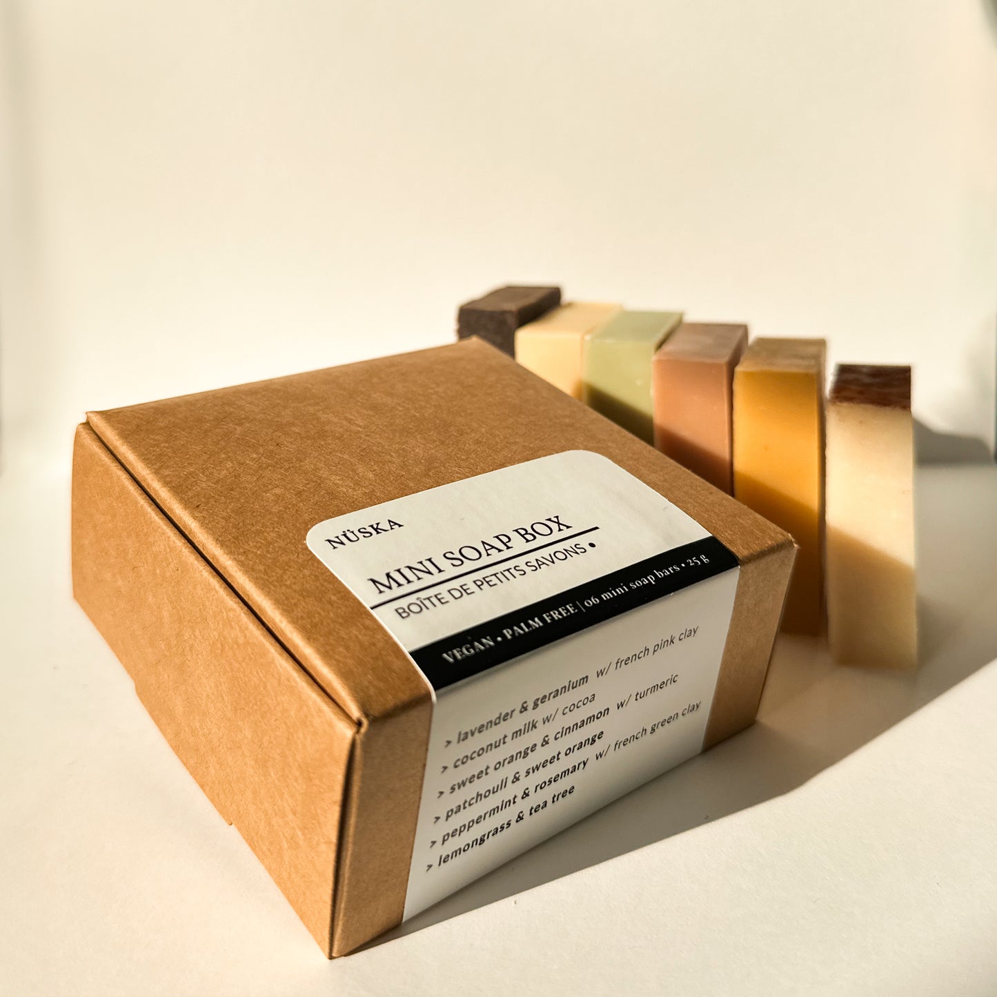 Mini Soap Box by NÜSKA, featuring six natural, handmade mini soap bars displayed next to a recyclable brown box with a label listing the soap scents. The soaps include a variety of colours, highlighting their natural ingredients. The box is labelled 'Vegan, Palm Free, 6 x 25g mini soap bars,' making it a perfect eco-friendly gift or self-care sampler.