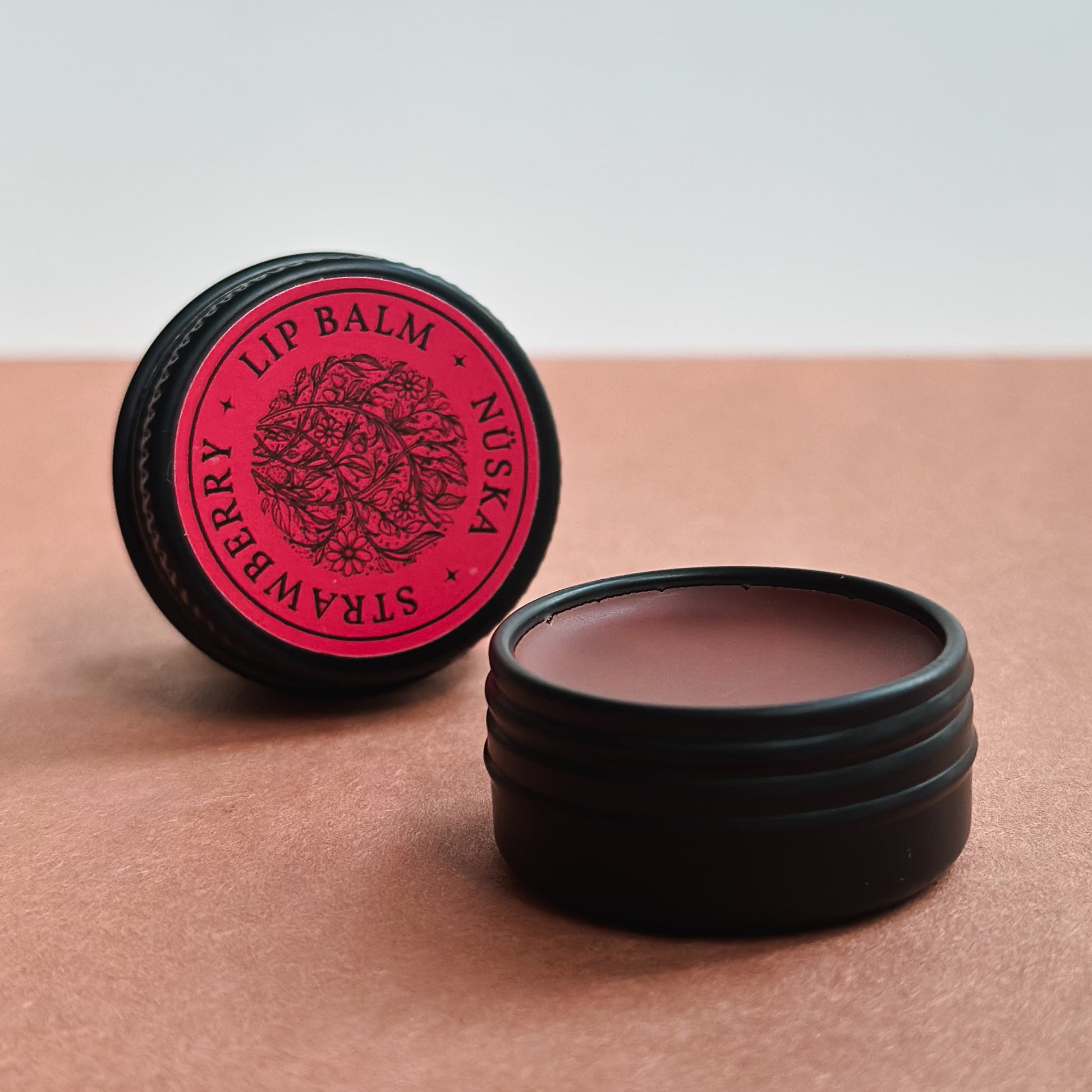 A black aluminum tin with a red label of Nuska strawberry natural tinted lip balm, and another open tin showing the colour and texture of the lip balm.