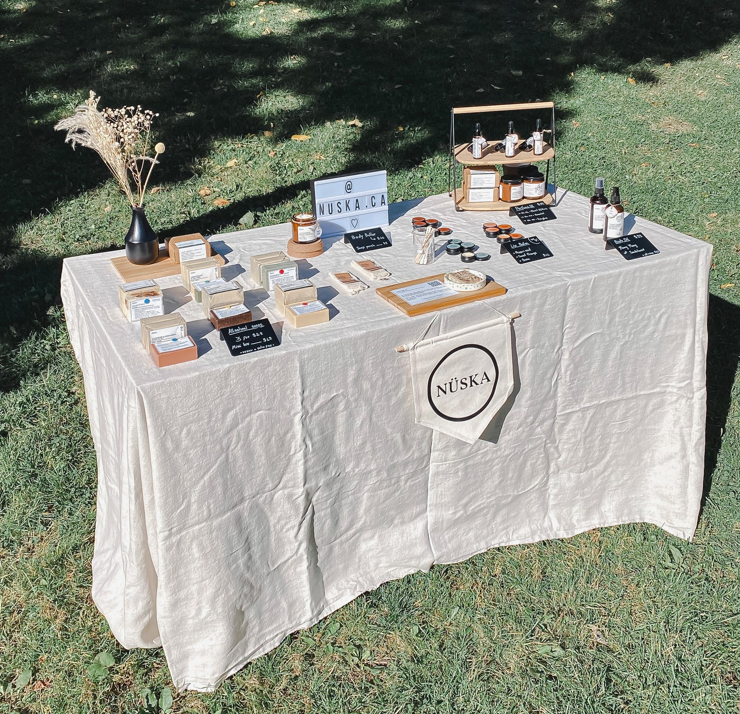 NÜSKA Conscious Cosmetics summer market table display featuring natural soap bars, lip balms, body oils, and body butters. Set outdoors on a grassy lawn, the minimalist setup includes product signage and a fabric banner with the NÜSKA logo