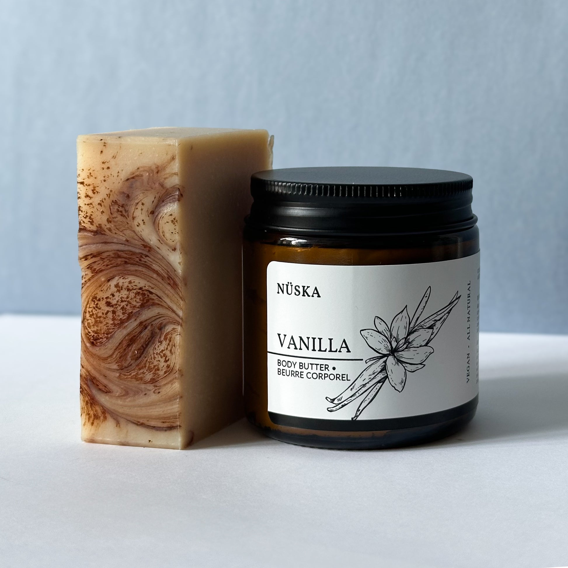 Amber glass jar of Nüska Vanilla Body Butter next to an unscented coconut natural moisturizer soap bar with a gentle swirl pattern, placed against a light background.
