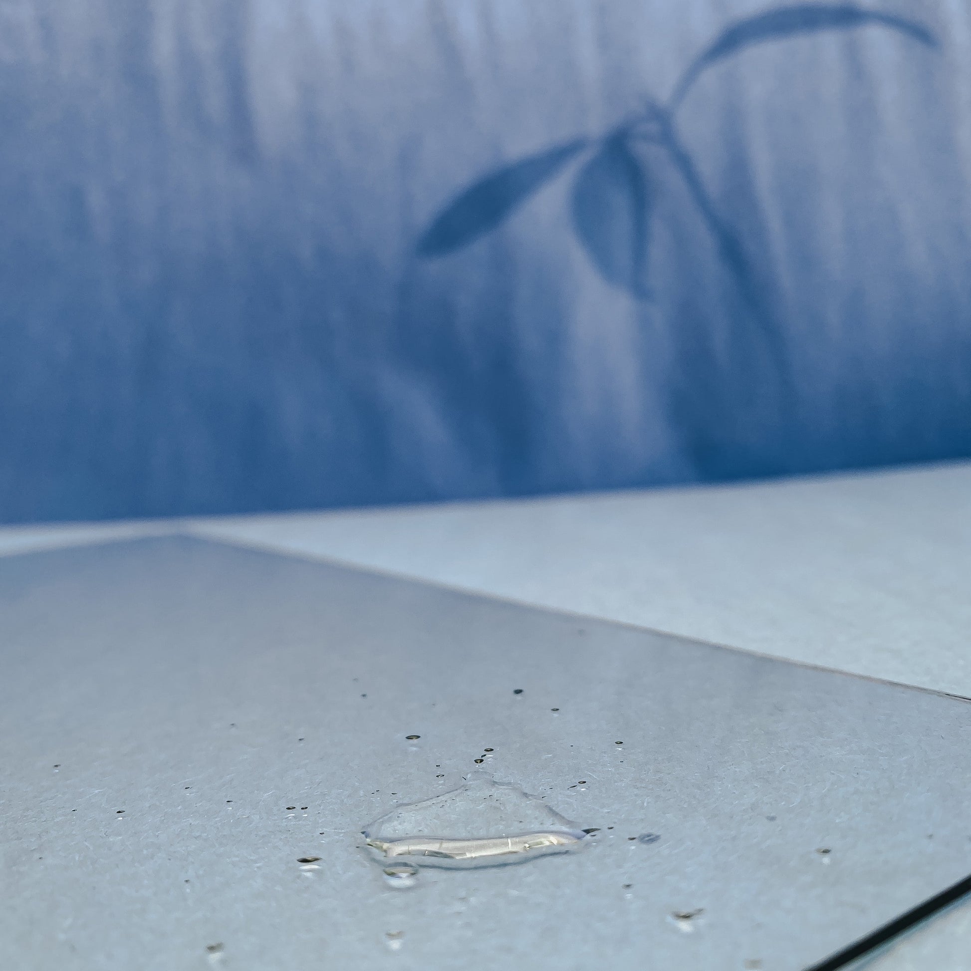 A drop of Nuska Ylang Ylang & Sandalwood Botanical body oil in a glass to show the texture, and a blue background.