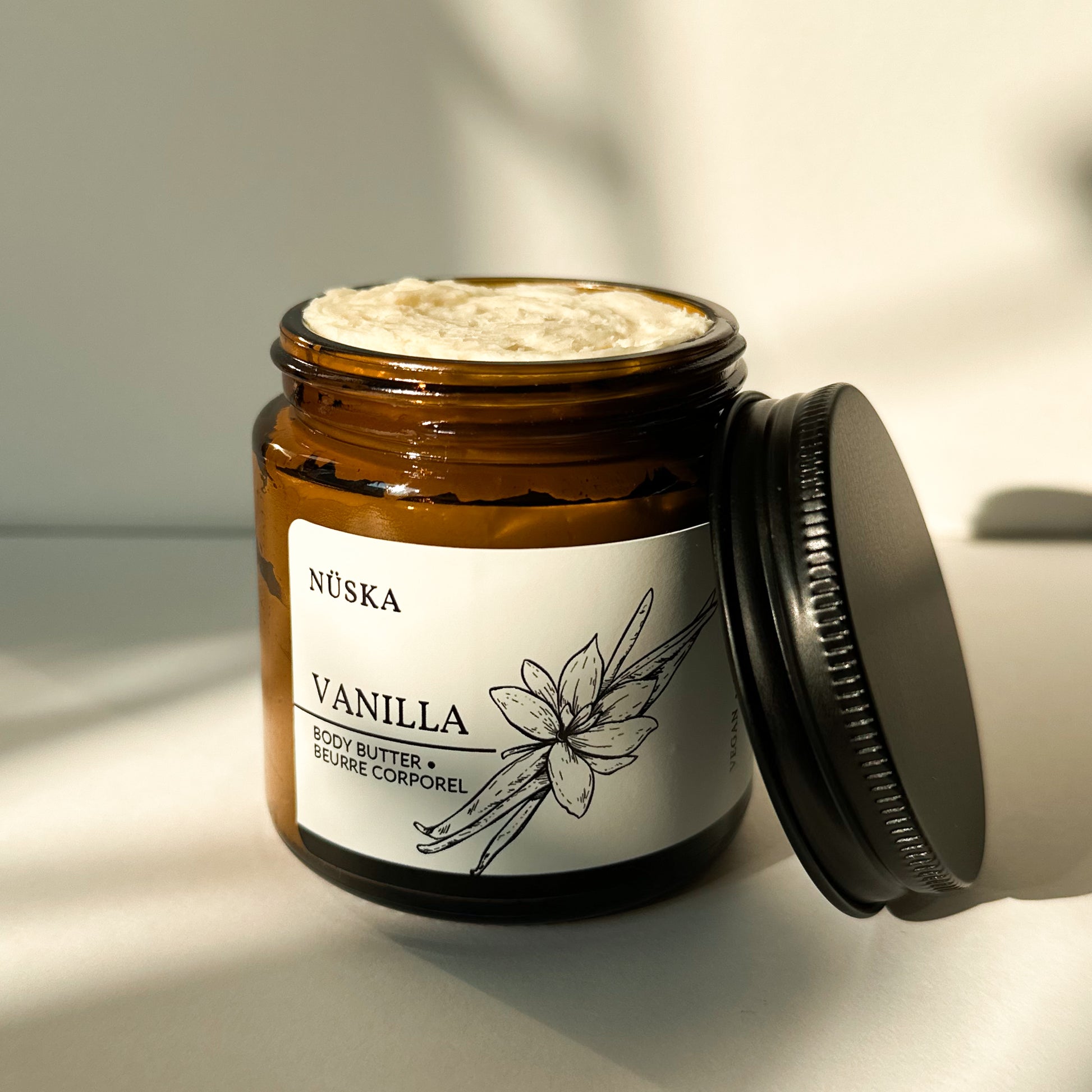 Nüska Vanilla Body Butter in an Open Amber Glass Jar with a Black Lid on the Side.