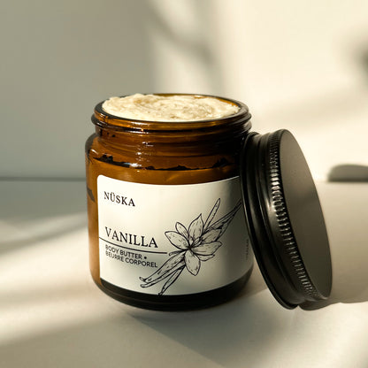 Nüska Vanilla Body Butter in an Open Amber Glass Jar with a Black Lid on the Side.