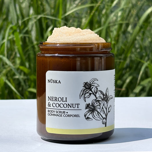 A opened glass jar of Nuska Neroli & Coconut Body Scrub showing the texture of the scrub with a green outdoor background.