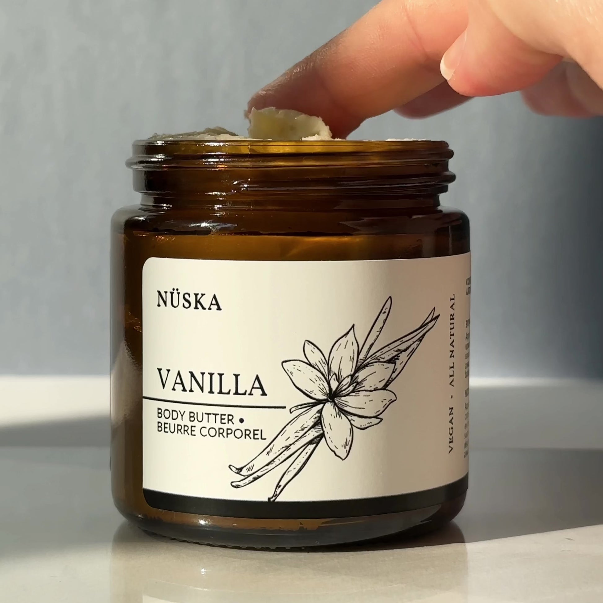 Hand picking up a small amount of Nüska Vanilla Body Butter from an amber jar, testing the creamy butter on fingertips as it gently melts into the skin, showing its smooth, moisturizing texture.