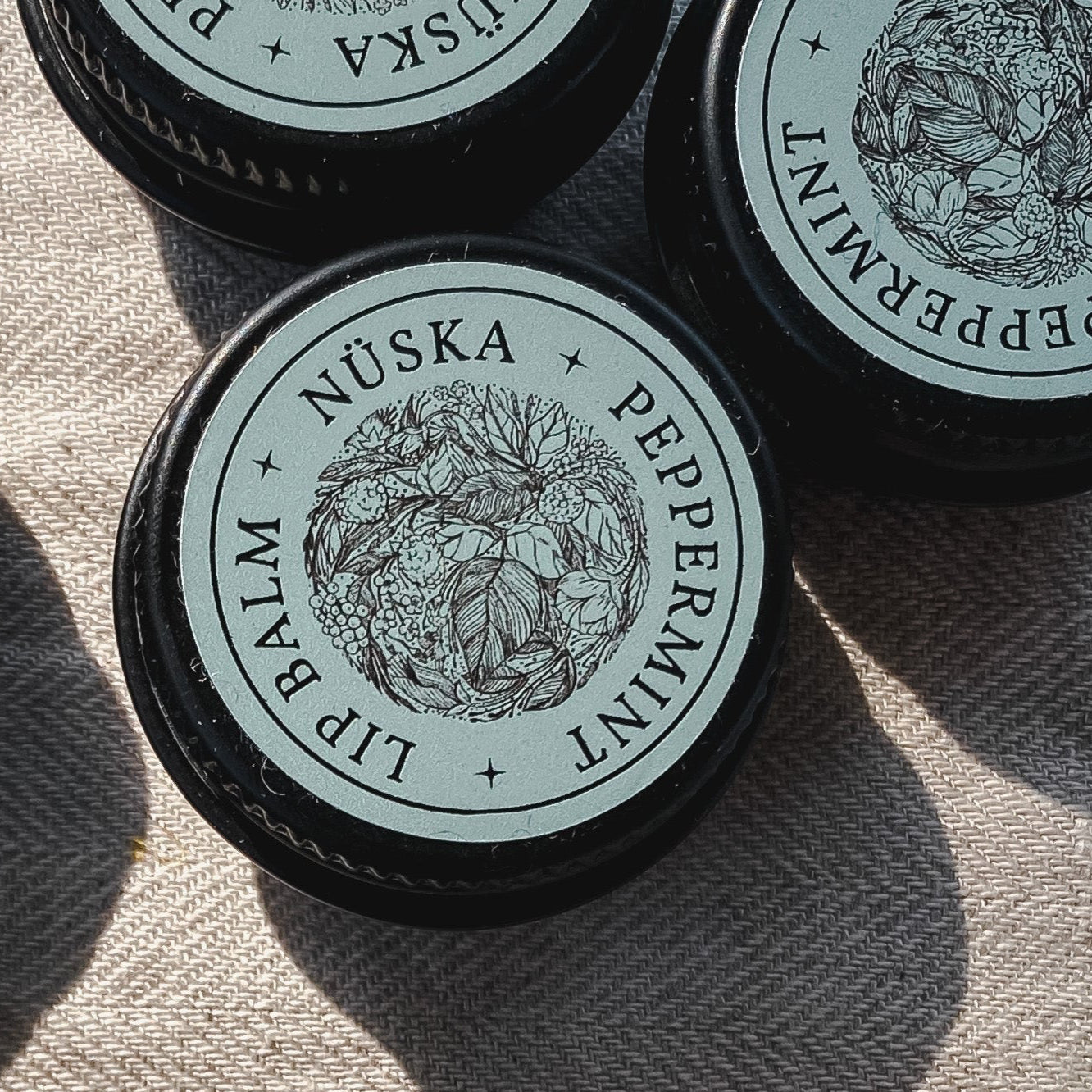 A black aluminum tin with a sage green label of Nuska peppermint natural lip balm.